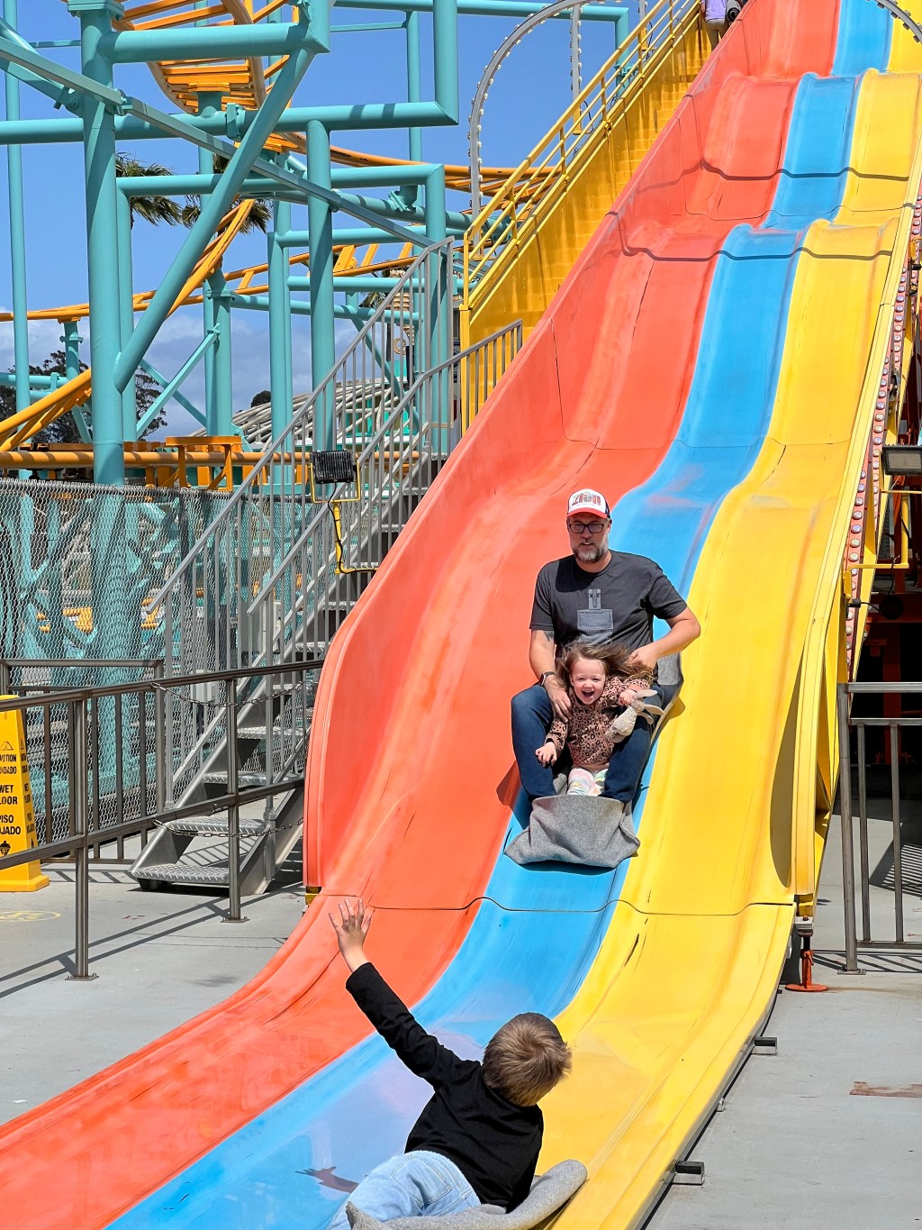 A man slides down giant slides with a child in his lap. Another child lays at the bottom with his hand raised, greeting them.